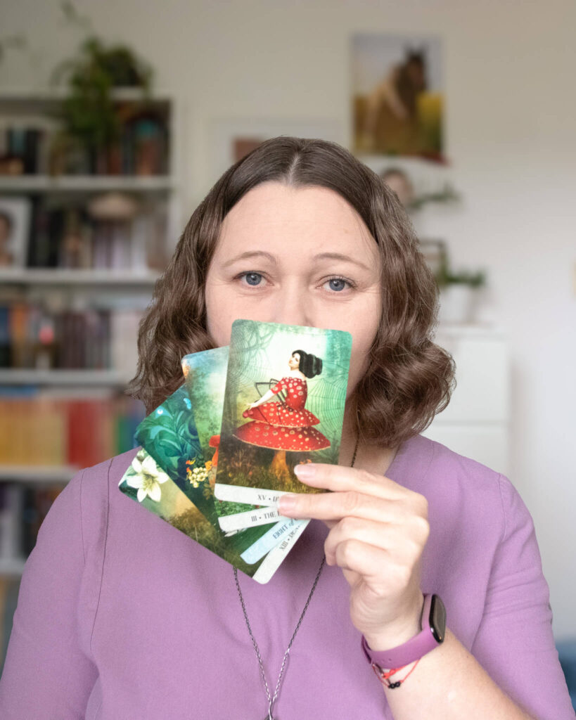 Autorka projektu Kompas Wiedźmy trzymająca karty tarota. Zdjęcie przedstawia kobietę w fioletowej koszuli, która z łagodnym spojrzeniem prezentuje karty, symbolizując intuicyjne prowadzenie i odczytywanie energii.